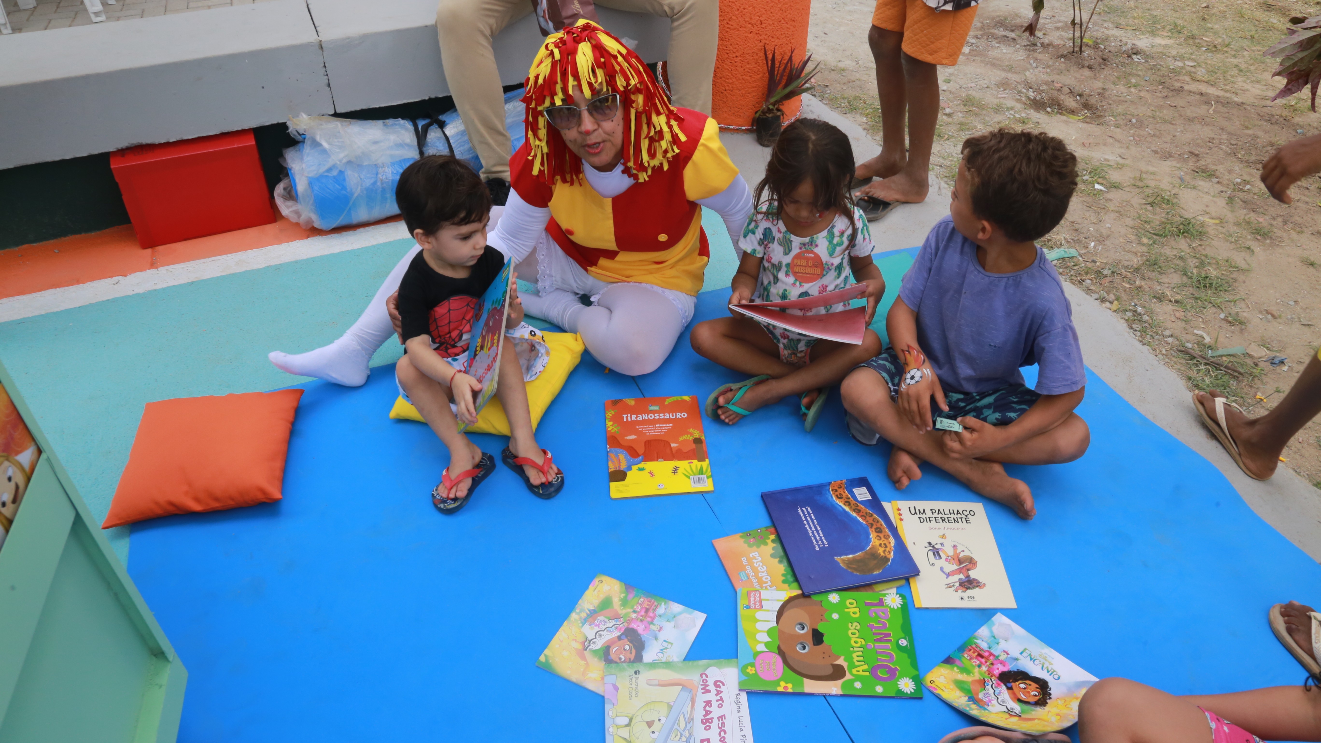 crianças lendo um livro em um tapete colocado na praça, acompanhados por facilitadora vestida de emília do sítio do pica-pau amarelo